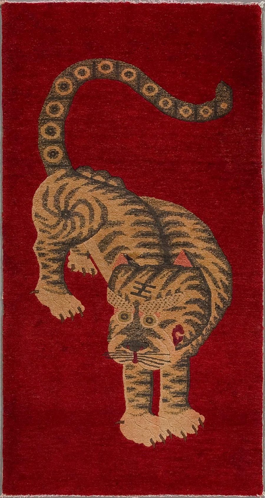Antiker Teppich mit stilisierter Darstellung eines Tigers auf rotem Hintergrund, der nach unten schaut, mit sichtbaren Streifen und Ringen auf dem Schwanz.