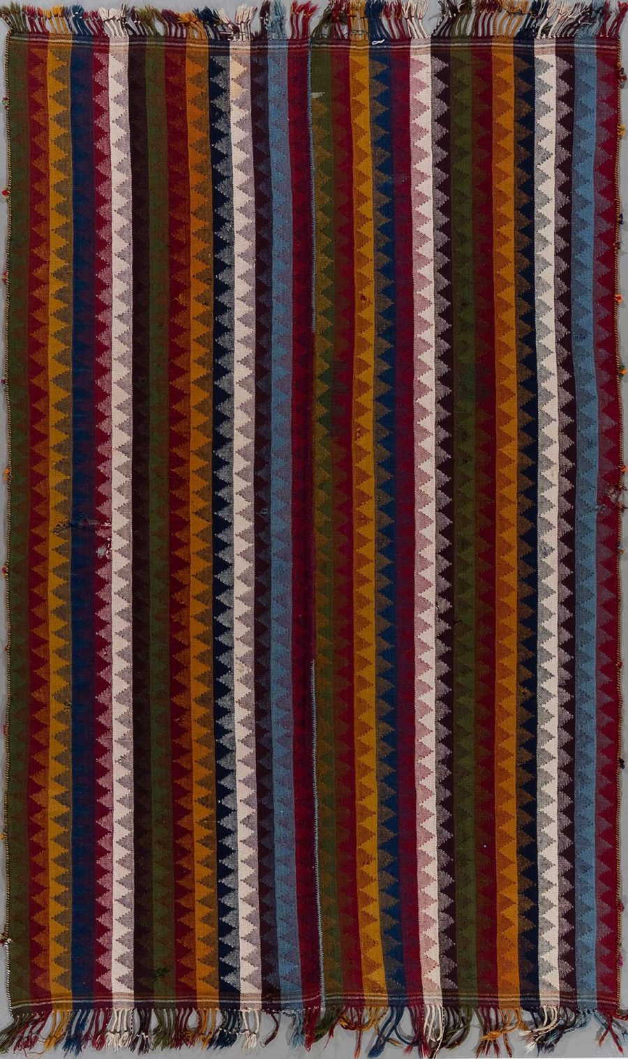 Vertikales Bild eines traditionellen, handgewebten Teppichs mit einem Muster aus mehrfarbigen vertikalen Streifen und gezackten Linien in verschiedenen Schattierungen von Rot, Blau, Braun und Weiß. Der Teppich hat Fransen an beiden Enden.