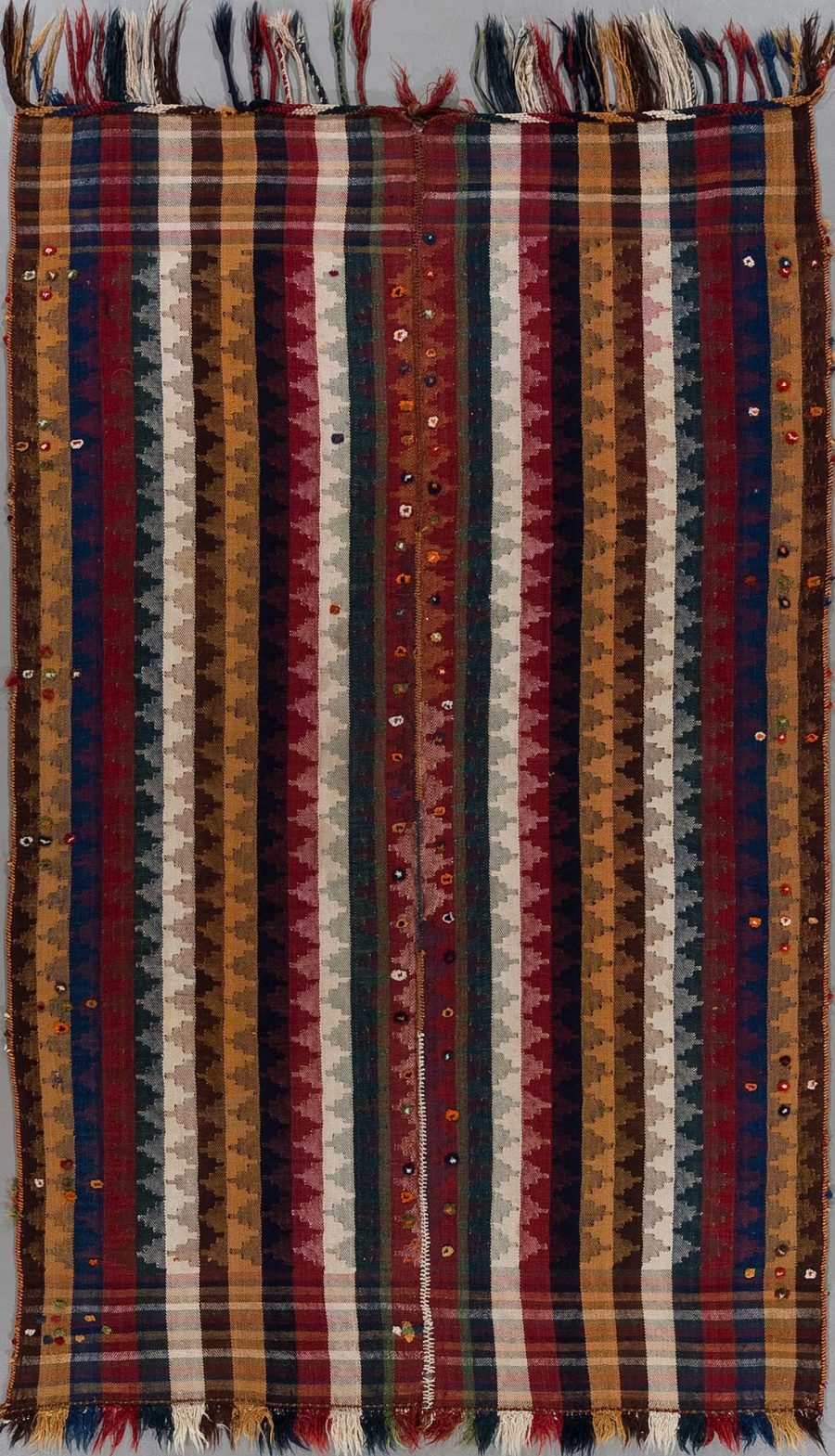Handgewebter Teppich mit Längsstreifen in verschiedenen Farben und Mustern, verziert mit mehrfarbigen Fransen an einem Ende und eingenähten kleinen Verzierungen über die gesamte Fläche verteilt.