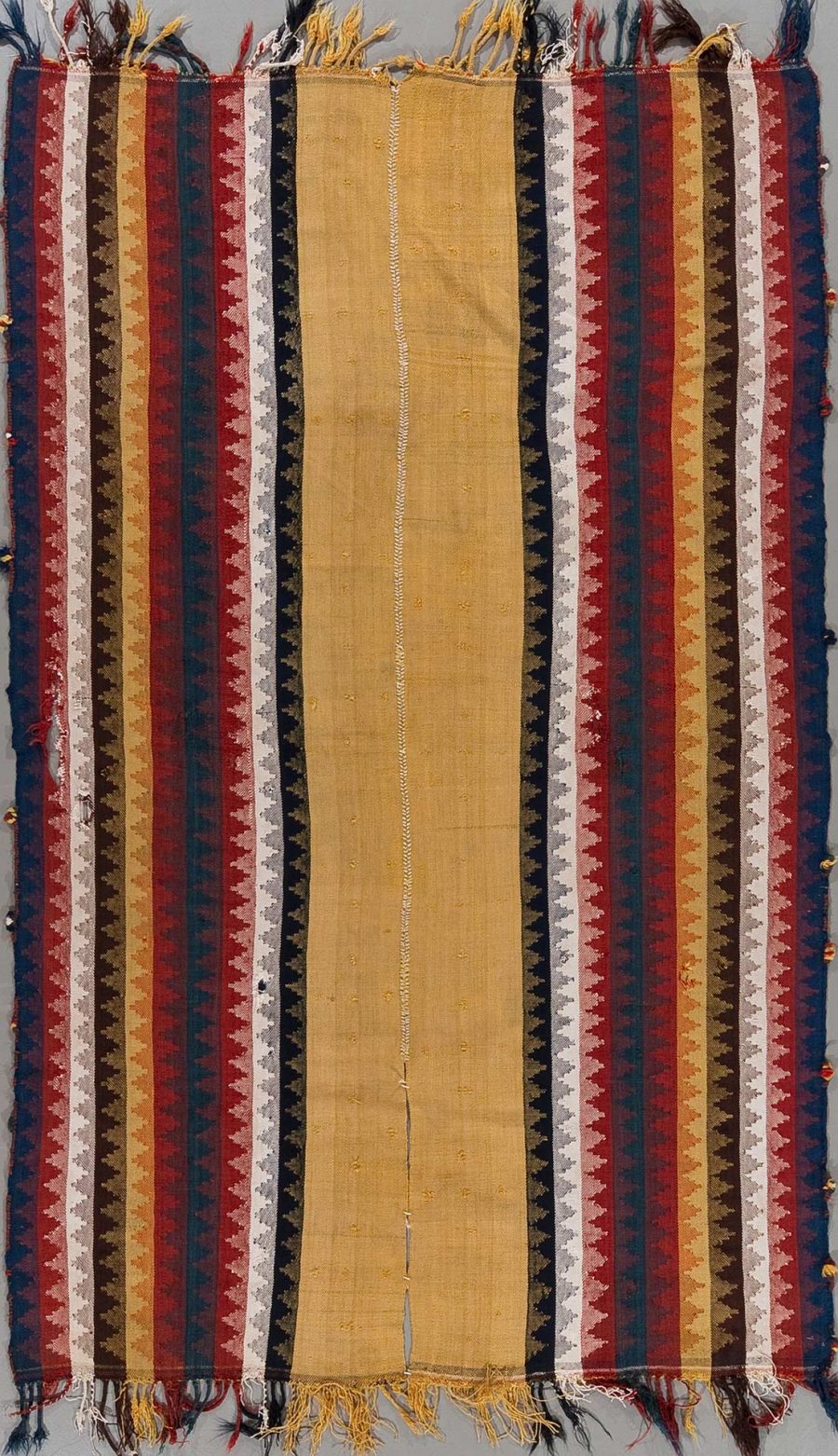 Vertikales Bild eines gewebten traditionellen Teppichs mit vielfarbigen Streifen und Zackenmuster sowie Franzen an den Enden.