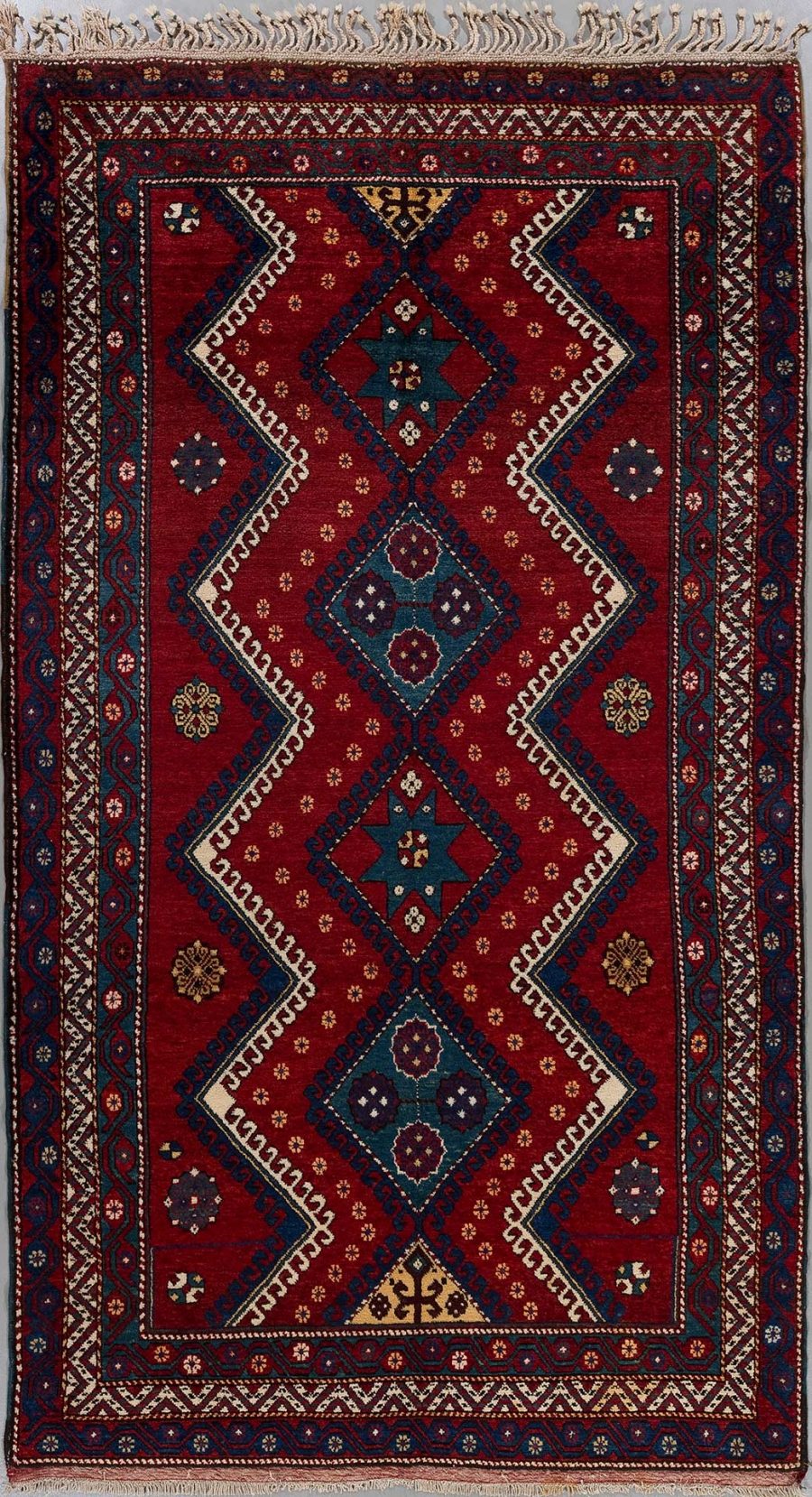Traditioneller handgeknüpfter Teppich mit einem komplexen Muster aus geometrischen Formen und Bordüren in Rot-, Blau- und Beigetönen.