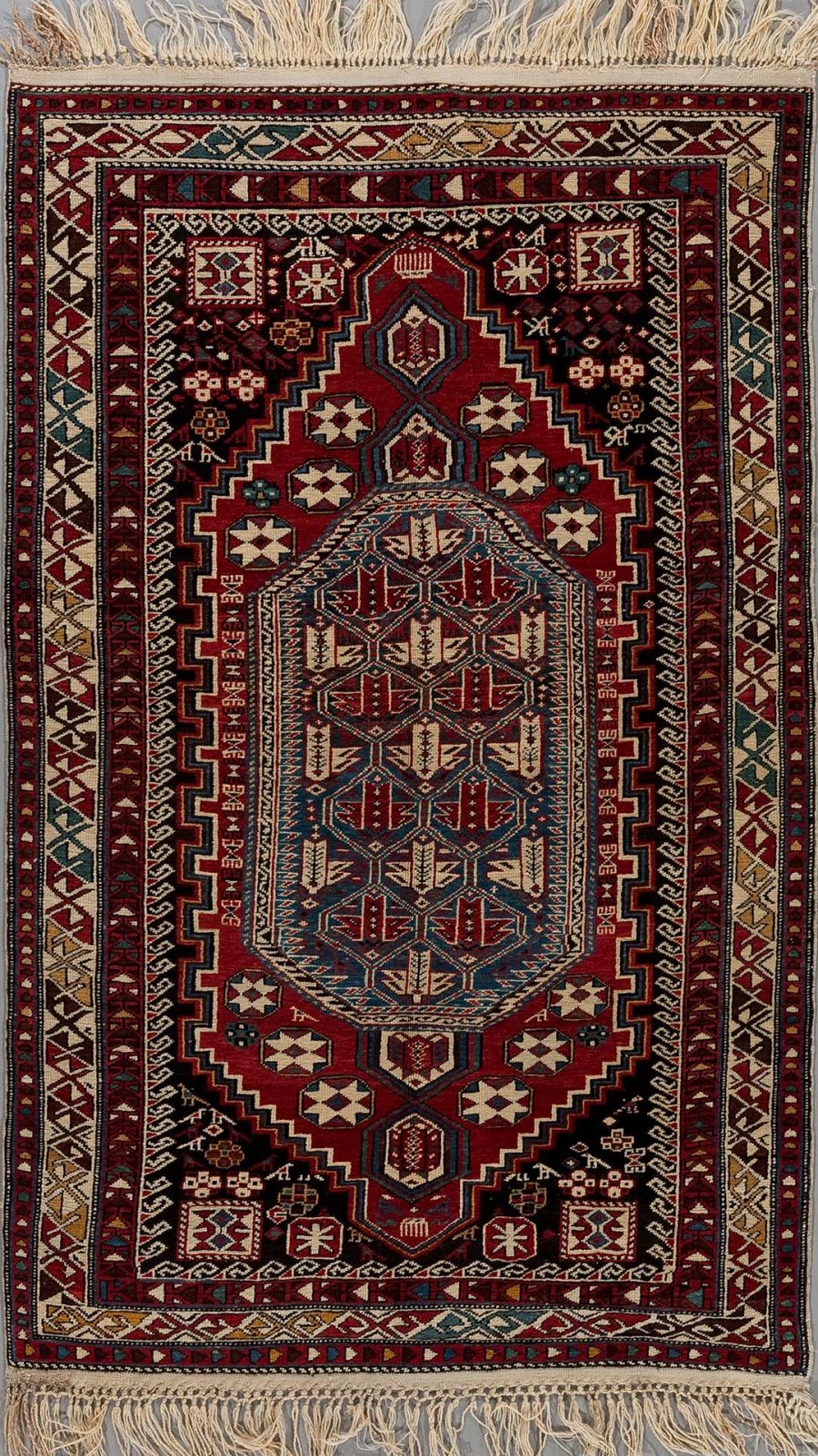 Detailaufnahme eines traditionellen handgeknüpften Teppichs mit komplexem Muster in Rot-, Blau-, Beige- und Schwarztönen, mit Fransen an den Enden.