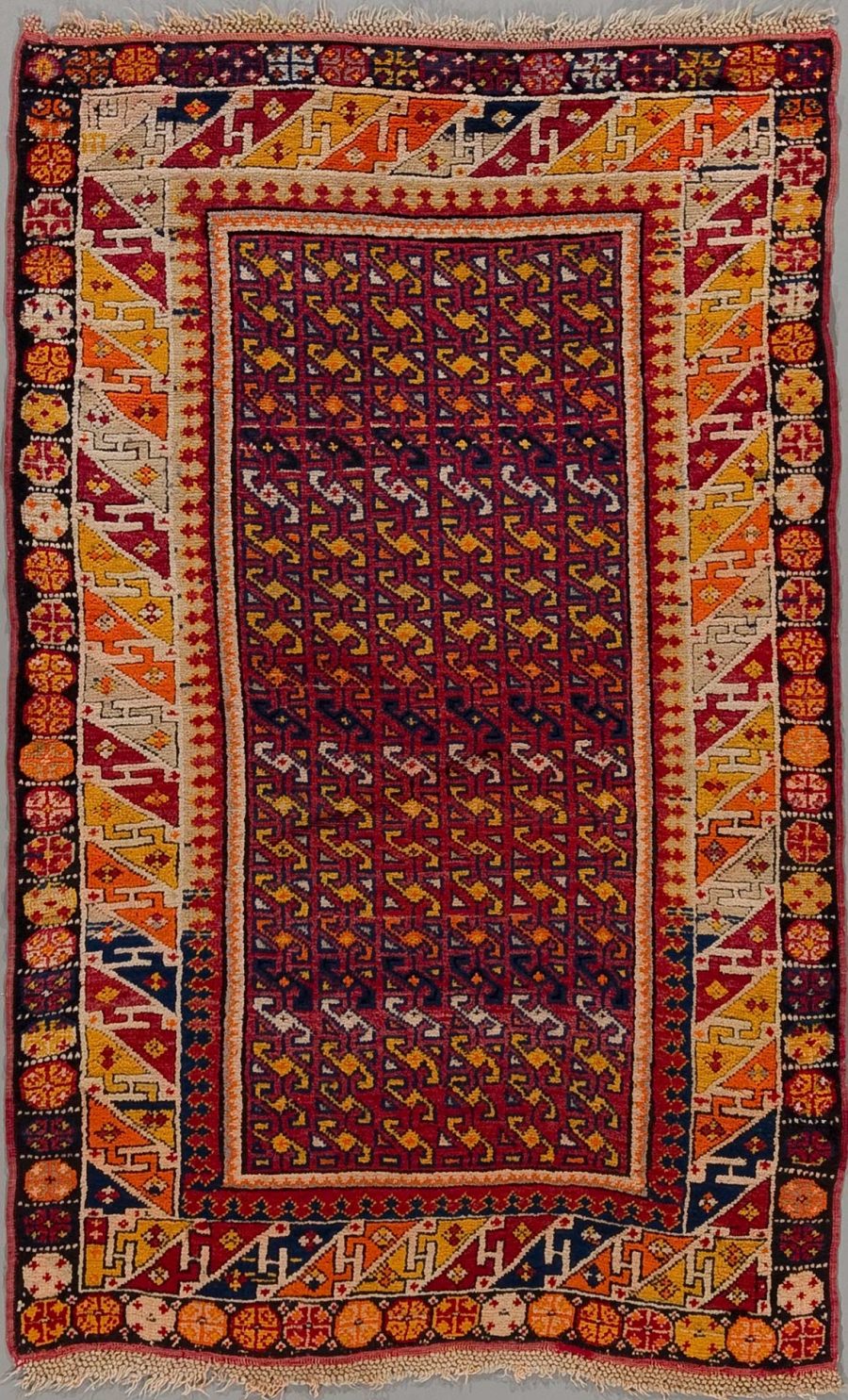 Detailreich gewebter, traditioneller Teppich mit komplexem Muster in vorwiegend roten, blauen und orangefarbenen Tönen, umrandet von einem dekorativen Rahmen mit geometrischen Formen und floralen Motiven.