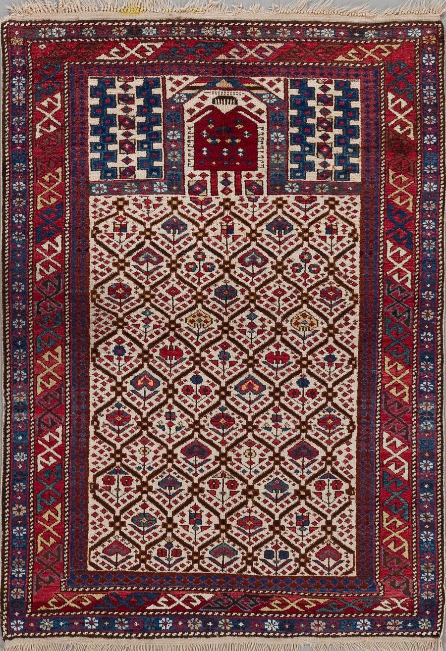 Handgeknüpfter Teppich mit detailliertem geometrischem Muster in verschiedenen Rottönen, umgeben von mehreren dekorativen Bordüren mit blauen, weißen und rotbraunen Akzenten. Oben auf dem Teppich ist ein kleines Torhaus-Motiv zu erkennen.