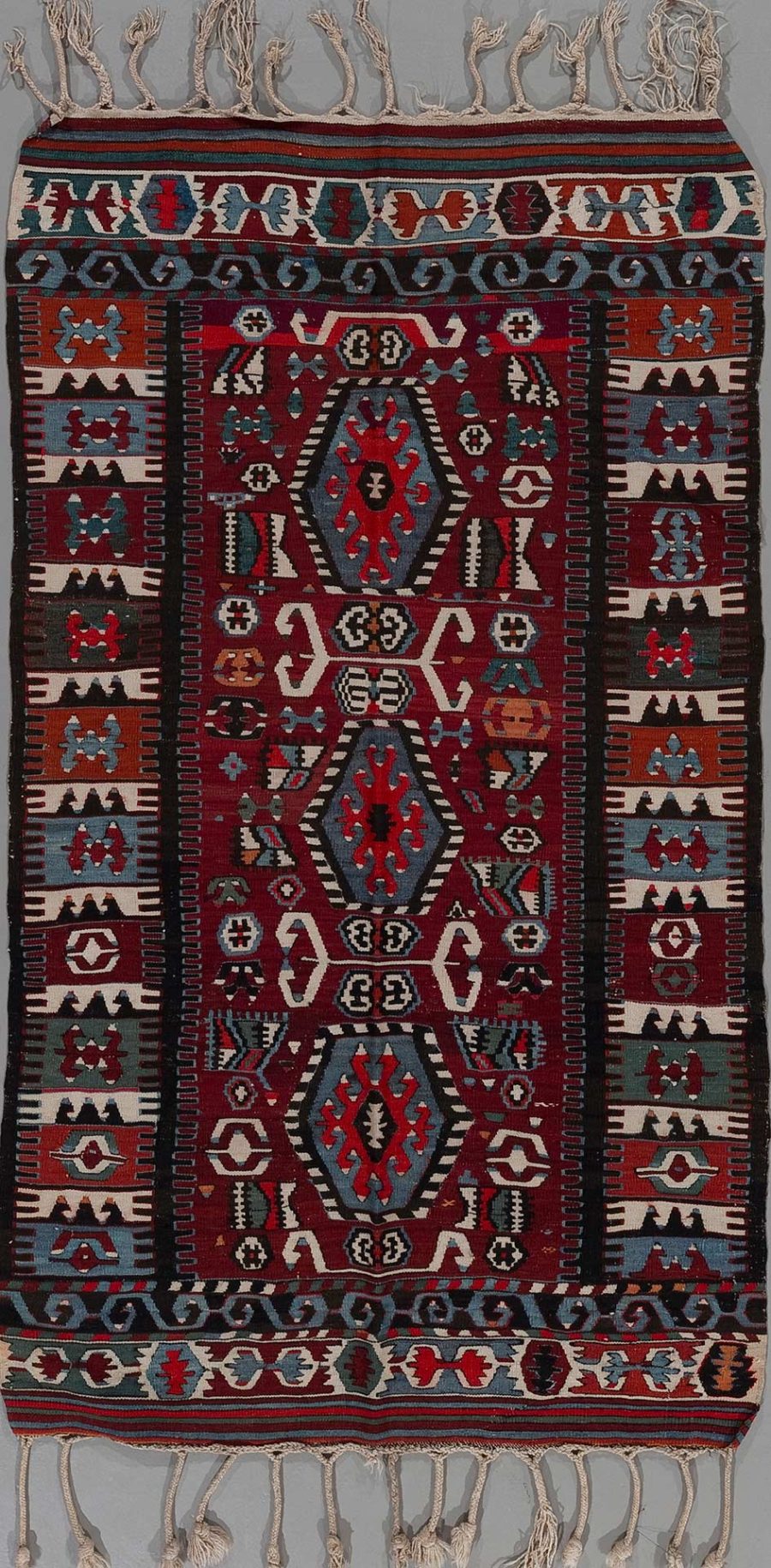 Traditioneller handgewebter Teppich mit komplizierten Mustern in Rot, Schwarz, Weiß und Blautönen, versehen mit Fransen an den Enden.