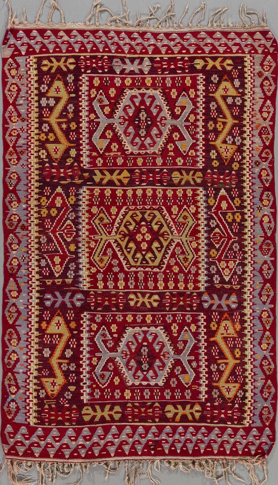 Traditioneller Teppich mit komplexem Muster in Rot-, Beige- und Gelbtönen sowie grauem Hintergrund, Fransen an den Enden.