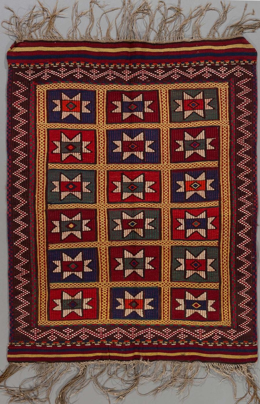 Traditioneller handgewebter Teppich mit Fransen und geometrischem Muster in Rot-, Blau-, Grün- und Beigetönen auf dunklem Grund.