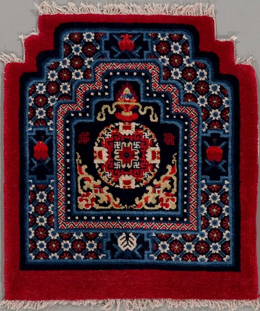 Traditioneller Teppich mit komplexem, geometrischem Muster in Rot-, Blau- und Beigetönen mit zentraler Raute und umgebenden Bordüren, aufgehängt an einer Wand.