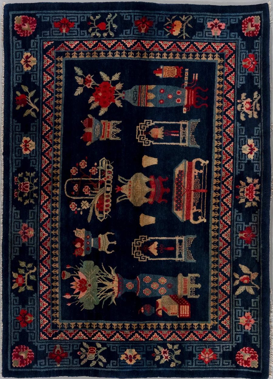 Traditioneller handgeknüpfter Teppich mit dunkelblauem Grund, umgeben von mehrfachen Bordüren in Grau und Beige, die geometrische Muster und rote Blüten zeigen. Im Zentrum sind mehrfarbige, stilisierte florale und tierähnliche Motive sowie Alltagsgegenstände angeordnet.