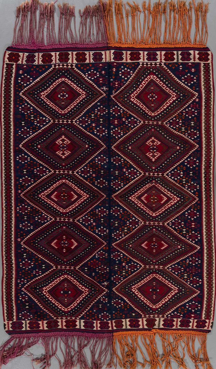 Traditioneller handgeknüpfter Teppich mit symmetrischem Muster in dunkelroten und marineblauen Farbtönen mit weißen und orangefarbenen Akzenten und Fransen an den Enden.