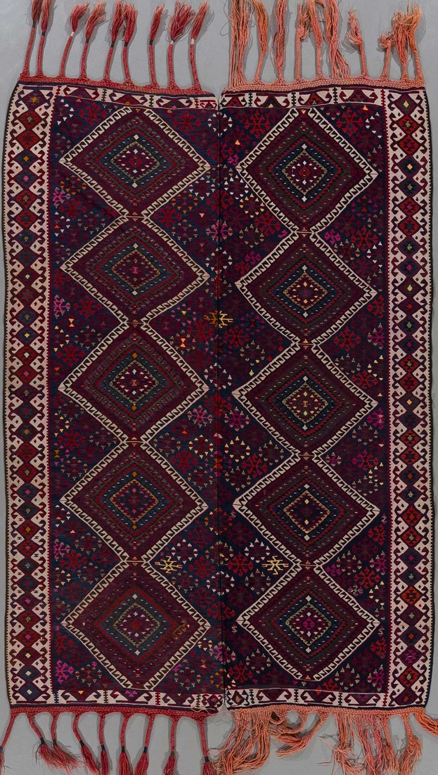 Traditioneller handgewebter Teppich mit geometrischem Muster in Dunkelrot, Schwarz und mehrfarbigen Akzenten, flankiert von verzierten Bordüren und Fransen an den Enden.