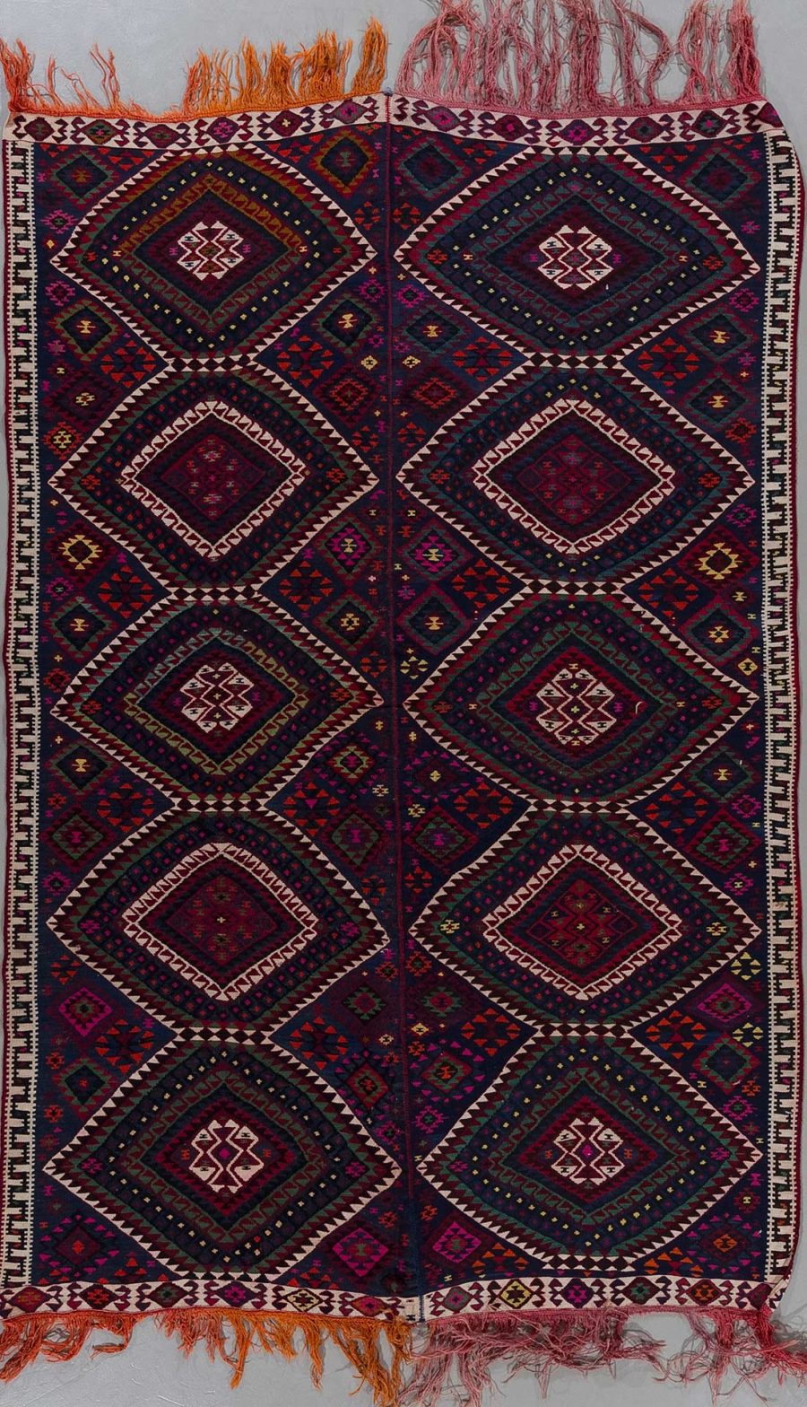 Handgewebter Teppich mit geometrischem Muster und Fransen, vornehmlich in dunkelroten und blauen Farbtönen, auf einem hellen Untergrund hängend.