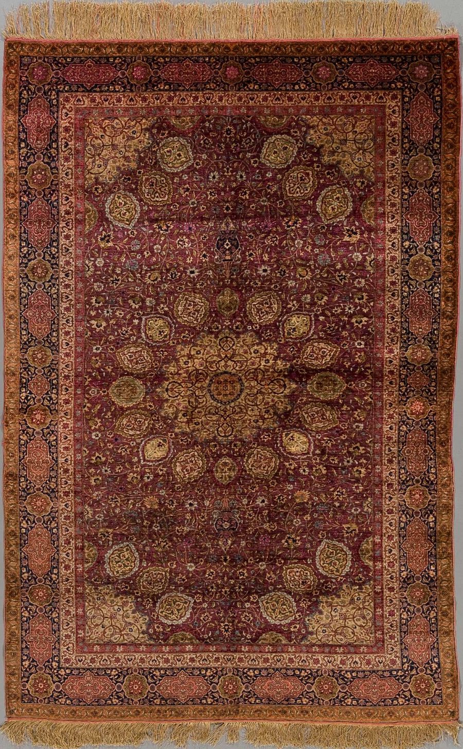 Antiker orientalischer Teppich mit dichtem Muster aus floralen und geometrischen Elementen in Rottönen mit Gold, Braun und blauen Akzenten, umgeben von mehreren Bordüren und mit Fransen an den Enden.