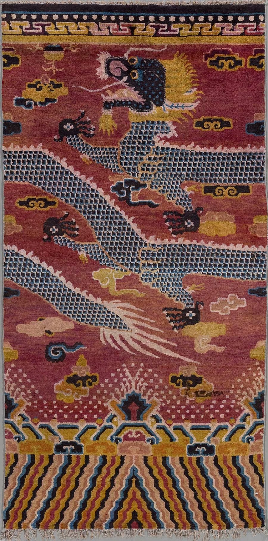 Alt Text: Handgeknüpfter Teppich mit traditionellen Mustern und Figuren, darunter ein zentrales Motiv, das an einen Drachen erinnert, auf einem bordeauxroten Hintergrund, umrahmt von geometrischen und abstrakten Formen in einer Vielzahl von Farben wie Gold, Blau, Rosa und Beige.