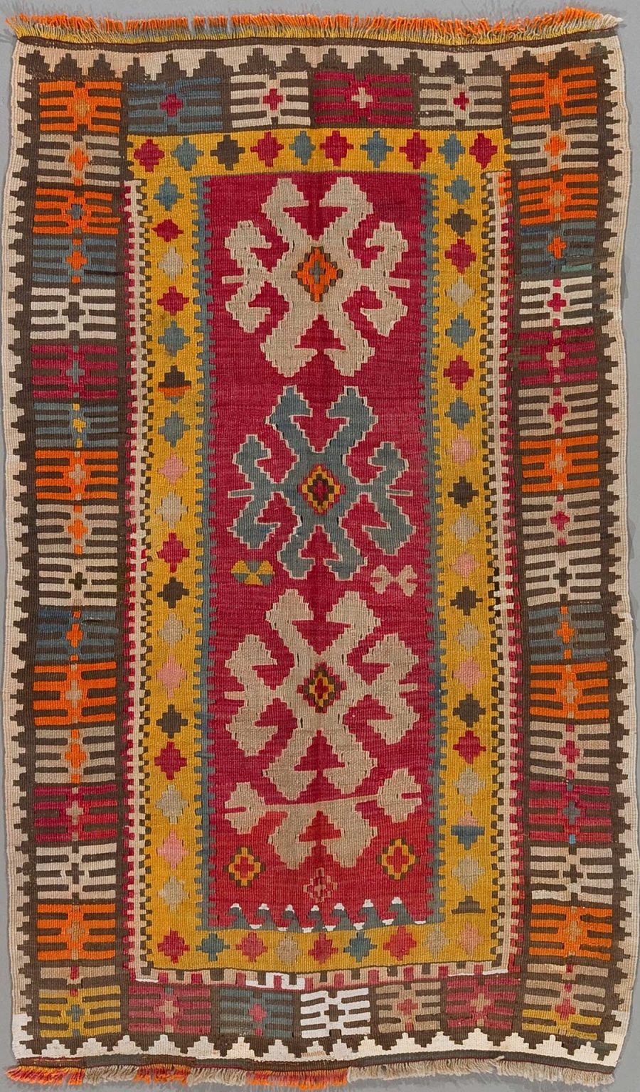 Handgewebter Teppich mit traditionellen Mustern in Rot, Gelb, Blau, Grau und Beige und zentralem Medaillon-Motiv, flankiert von dekorativen Bordüren und Fransen an den Enden.
