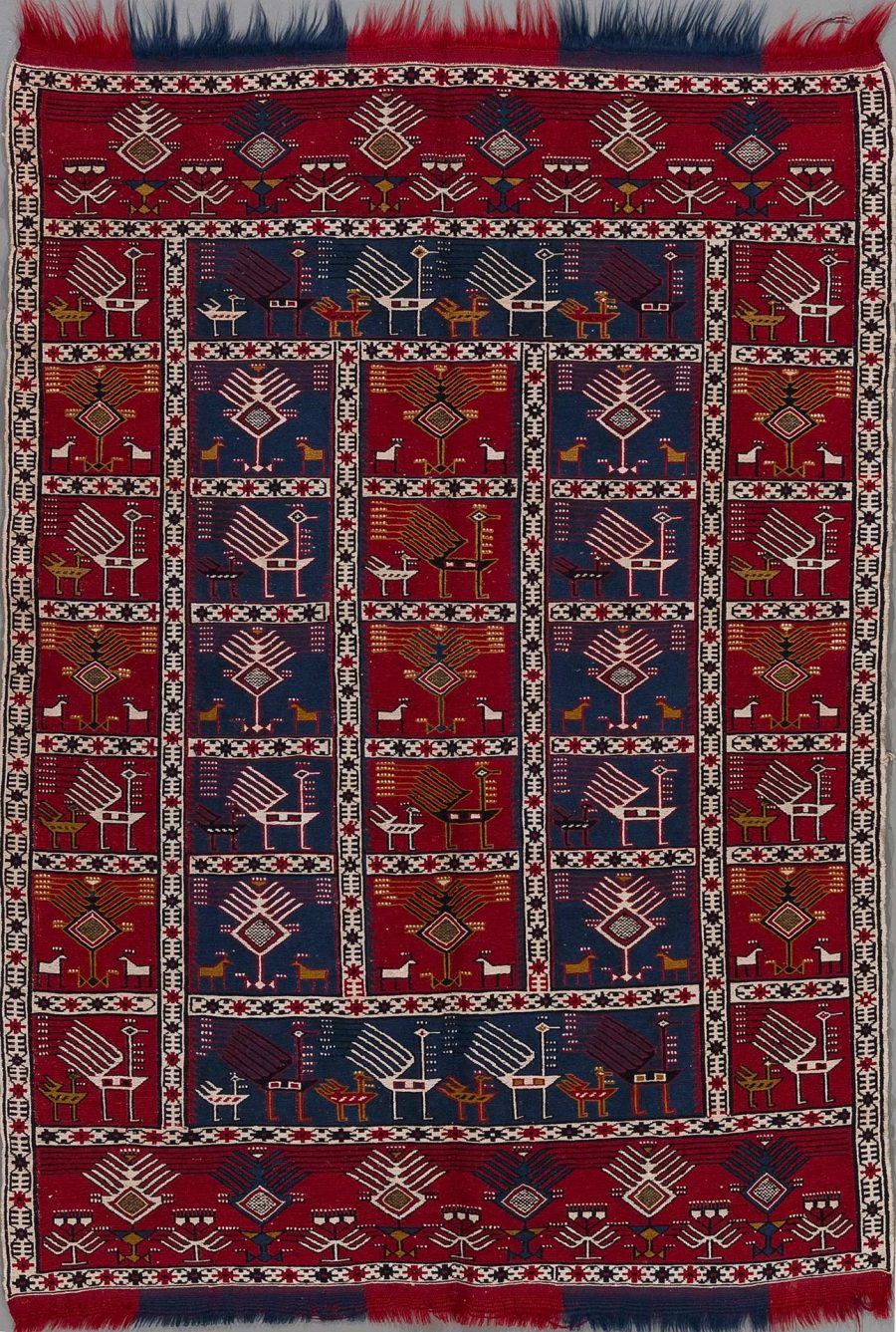 Traditioneller handgewebter Teppich mit komplexem Muster in Rot-, Blau- und Beigetönen, verziert mit geometrischen und stilisierten floralen Motiven sowie Tierdarstellungen, flankiert von dekorativen Bordüren und abgeschlossen mit Fransen an beiden Enden.