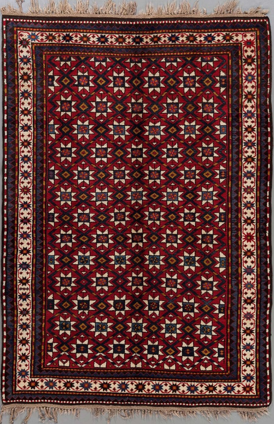 Traditioneller orientalischer Teppich mit komplexem geometrischem Muster in Rot, Blau und Beige-Tönen, umgeben von mehreren Bordüren und Fransen an den kurzen Enden.