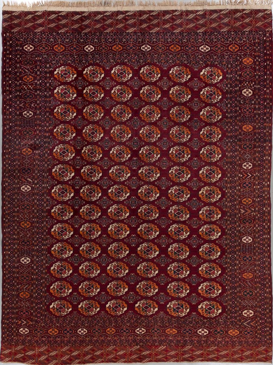 Traditioneller handgeknüpfter Teppich mit dichtem Muster aus wiederholenden geometrischen Formen und Ornamenten in Rottönen, umrahmt von mehreren Bordüren mit komplexen Mustern, aufgehängt an einer Wand zur Präsentation.