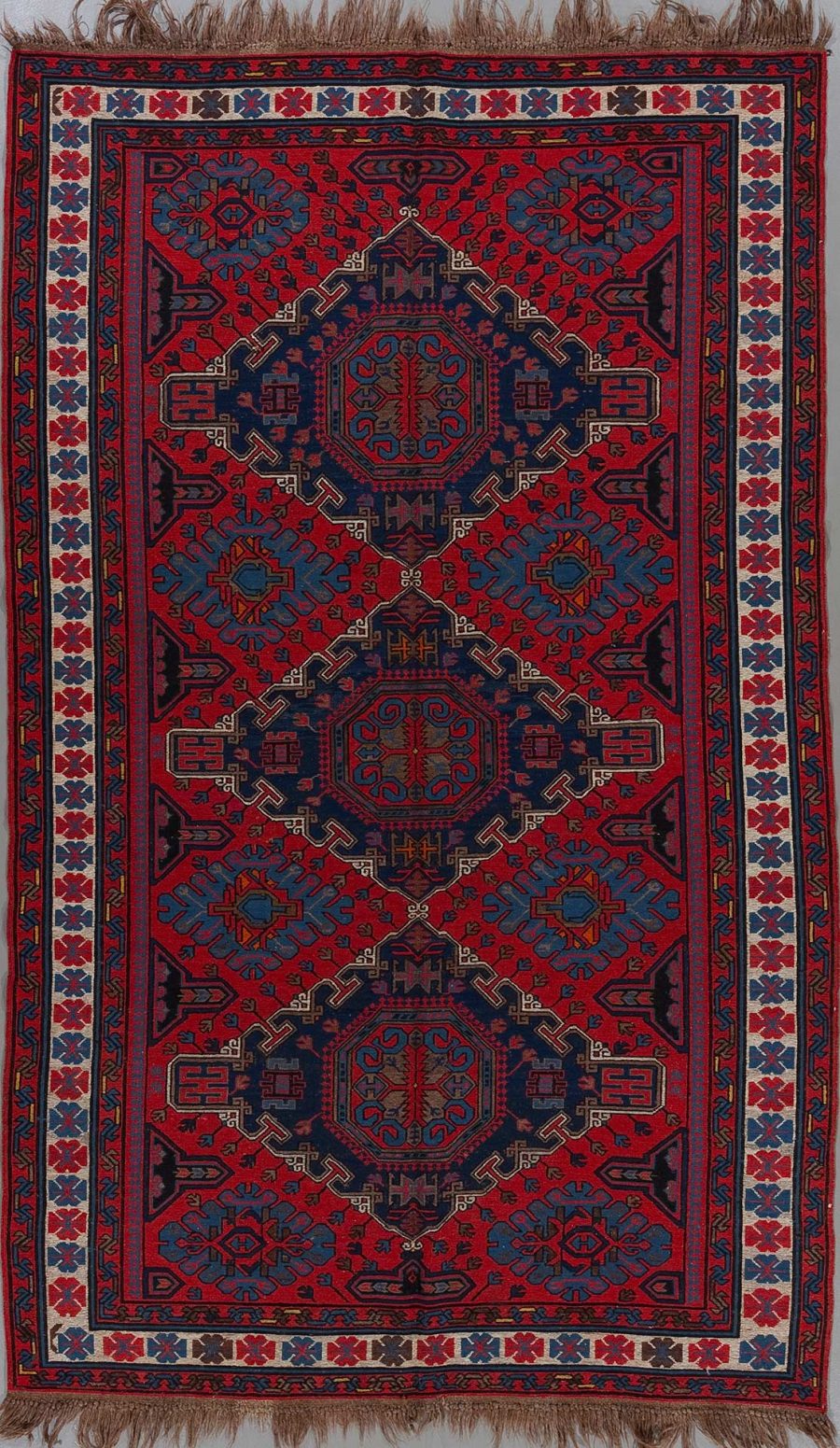Detailaufnahme eines traditionellen orientalischen Teppichs mit komplexem Muster aus geometrischen Formen und pflanzlichen Motiven in Rot-, Blau- und Beigetönen, eingefasst von mehreren Bordüren und verziert mit Fransen am oberen und unteren Ende.