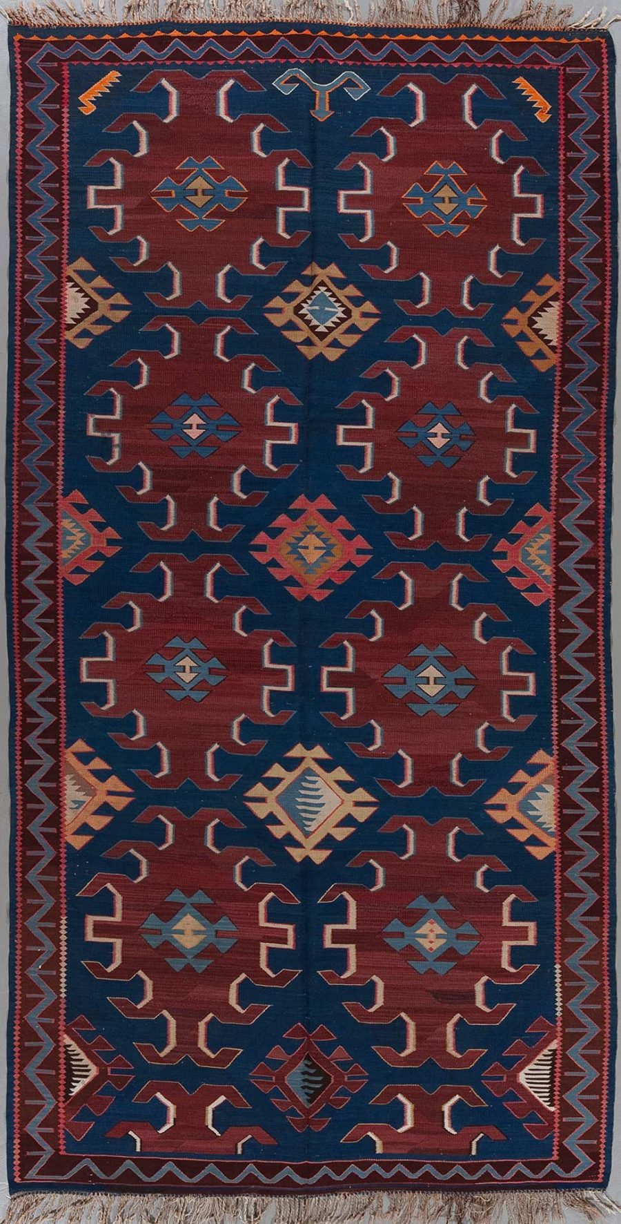 Handgeknüpfter Teppich mit traditionellem geometrischem Muster in Farbtönen von Blau, Rot, Beige und Orange, umgeben von dekorativen Bordüren und Fransen an beiden Enden.
