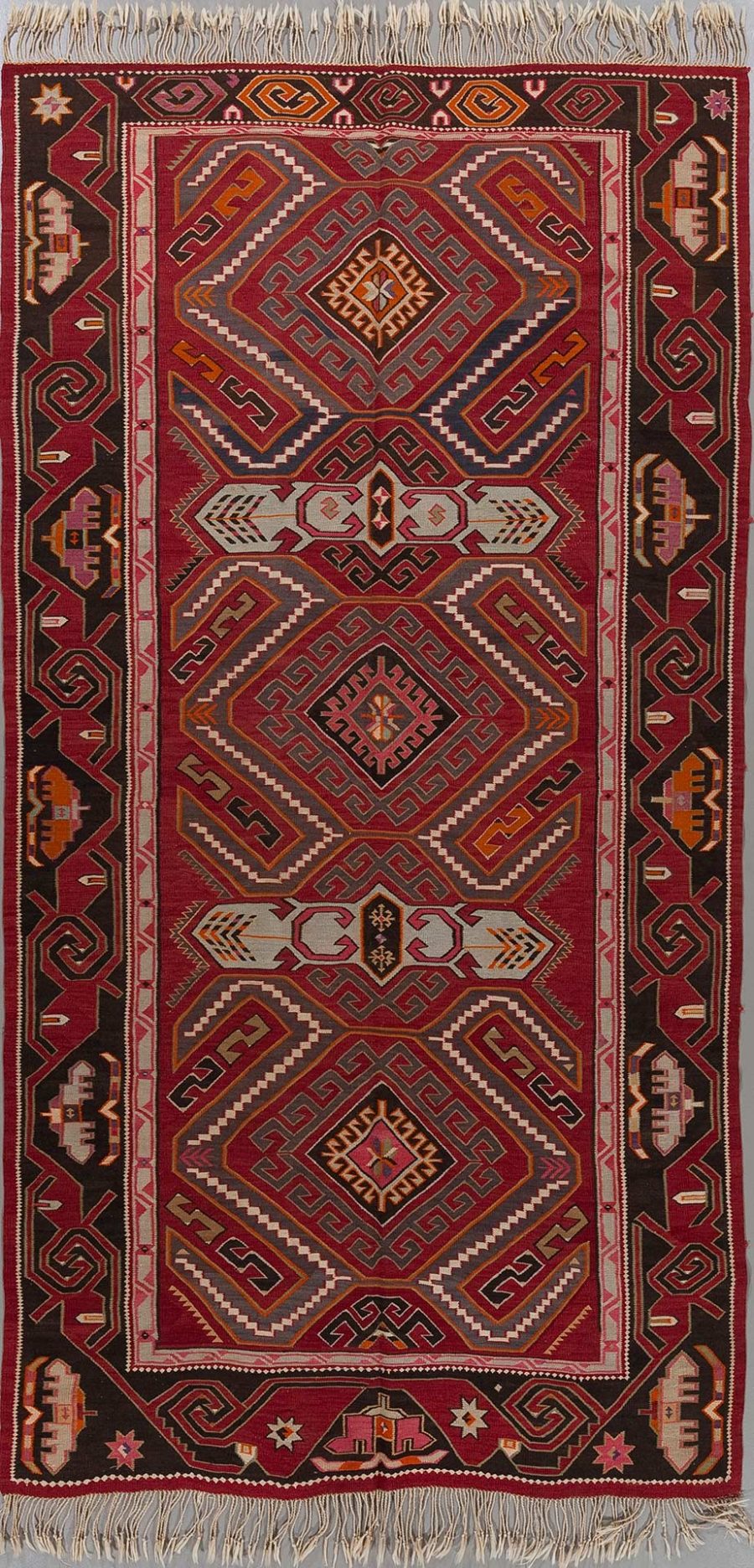 Traditioneller Teppich mit komplexem, geometrischem Muster in Rot-, Schwarz-, Weiß- und Orangetönen, umgeben von dekorativen Bordüren und Fransen an den Enden.