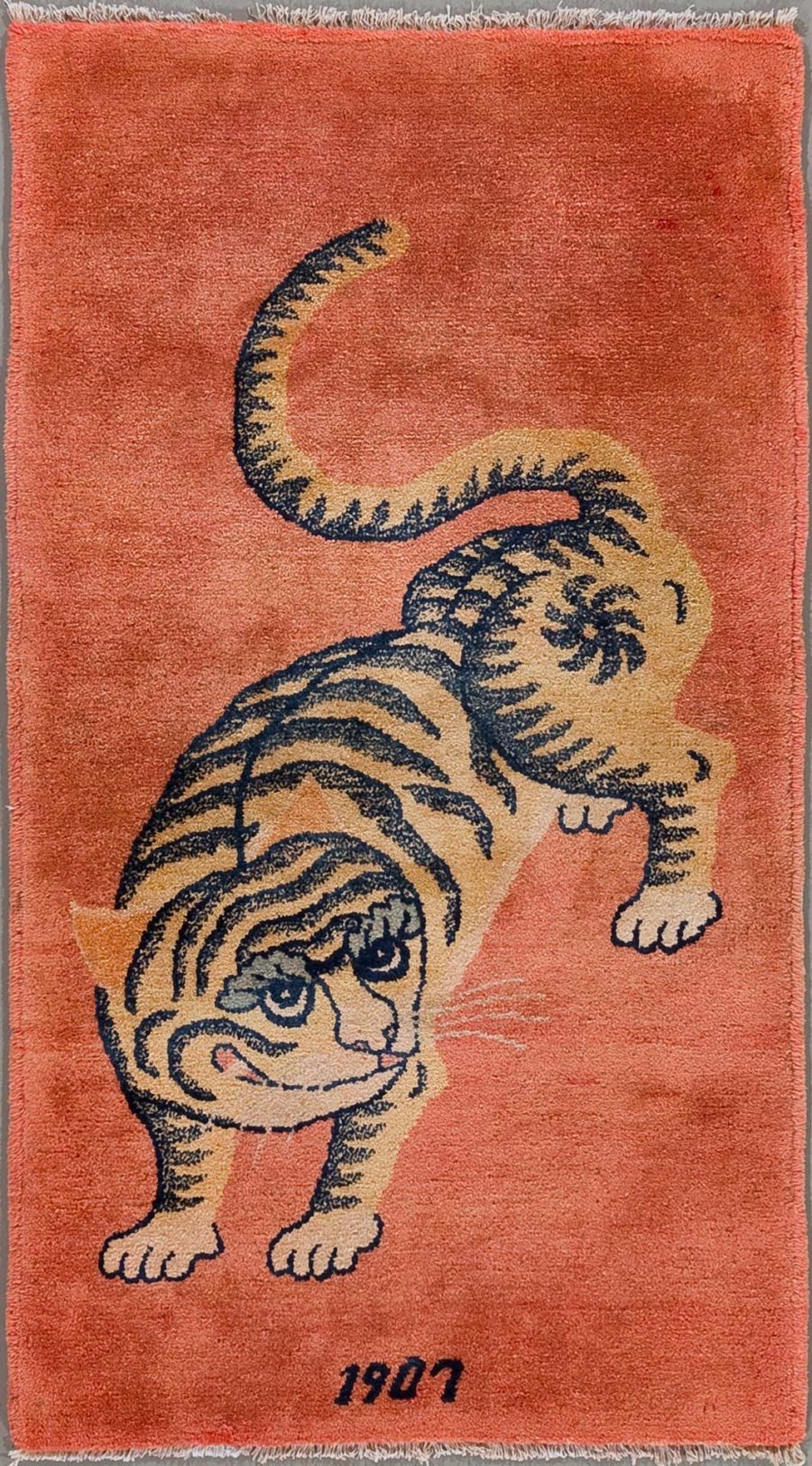 Vintage-Teppich mit einer stilisierten Darstellung eines Tigers auf orangefarbenem Grund, Jahreszahl 1907 im unteren Bereich.