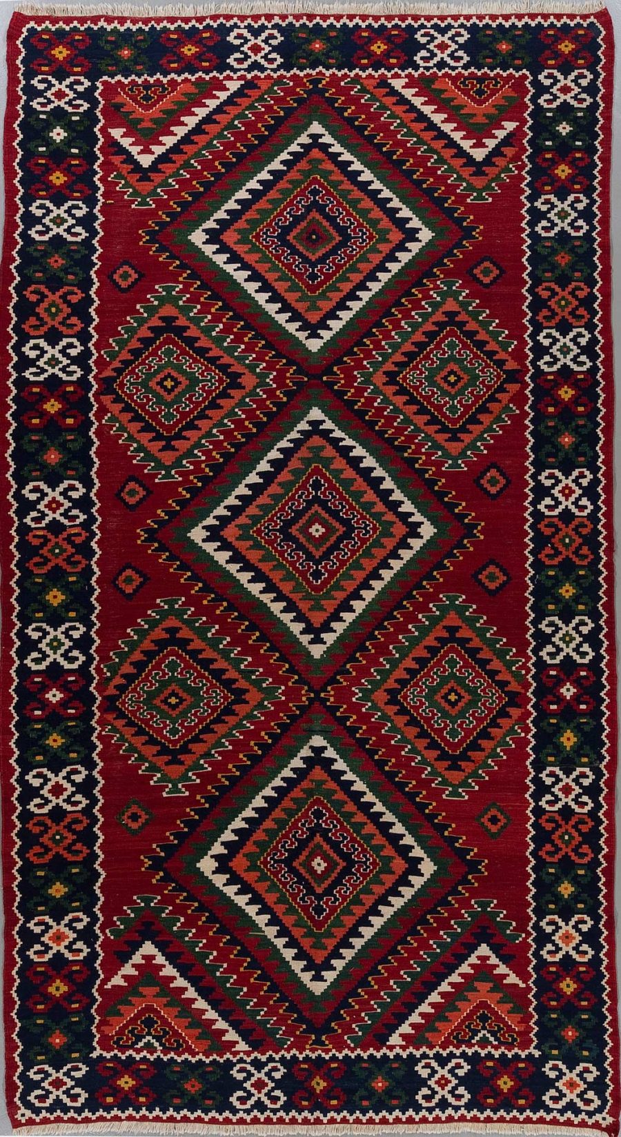 Traditioneller handgeknüpfter Teppich mit geometrischen Mustern und einem dominanten Rot, flankiert von grünen und beige Akzenten sowie Grenzdesigns in Dunkelblau und Weiß.