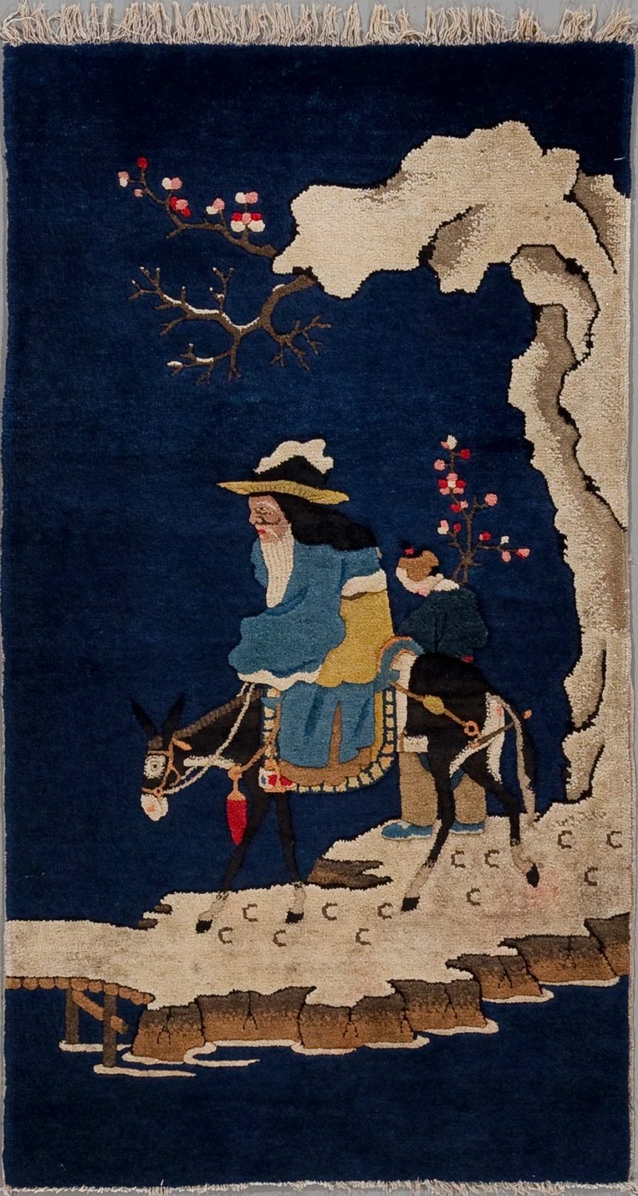 Handgewebter Teppich mit darstellender Szene: Ein älterer Reisender in traditioneller Kleidung sitzt auf einem Esel, der von einem jungen Begleiter geführt wird. Sie überqueren eine Brücke in einer nächtlichen Landschaft unter blühenden Bäumen und neben einem großen, knorrigen Baum, deren Konturen sich vom dunkelblauen Hintergrund abheben.