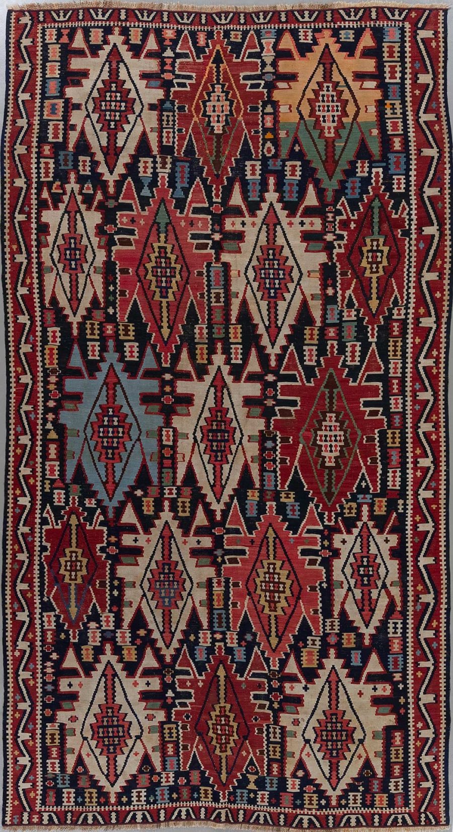 Ein handgewebter Teppich mit einem komplexen, symmetrischen Muster aus Diamanten und geometrischen Formen in Rot, Blau, Beige und Schwarztönen.