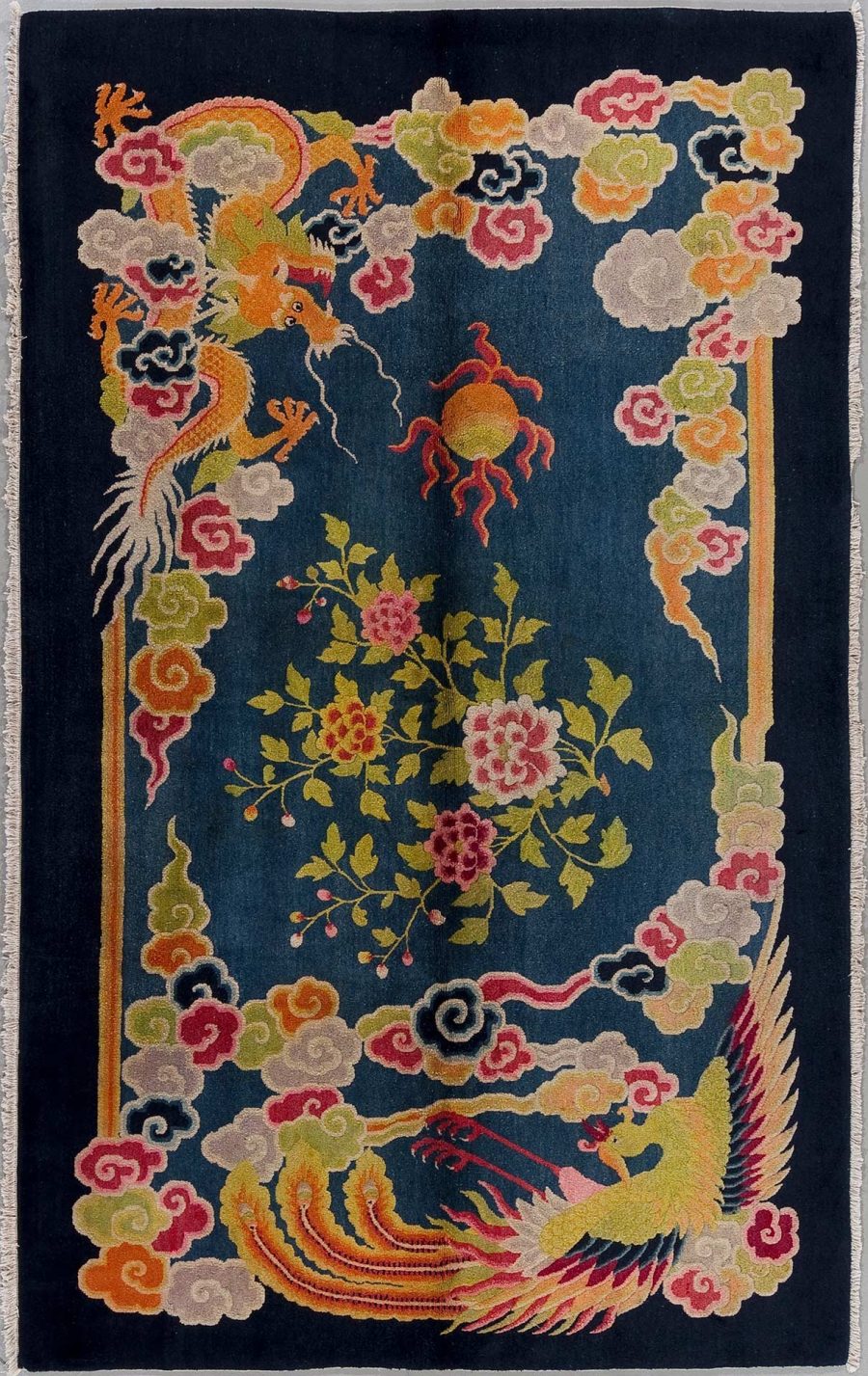 Antik wirkender, orientalischer Teppich mit dunkelblauem Grund, darauf sind ein roter Phoenix und ein orangefarbener Drache neben blühenden Pflanzen und Wolkenmotiven in lebendigen Farben abgebildet.