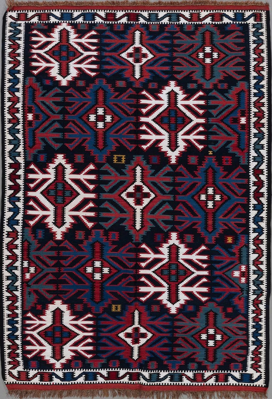 Handgewebter Teppich mit traditionellem Muster in Dunkelblau, Rot, Weiß und kleinen Akzenten in Gelb, symmetrische geometrische Designs und Rautenformen, umrandet von einem gezackten Muster, Fransen an einem Ende.