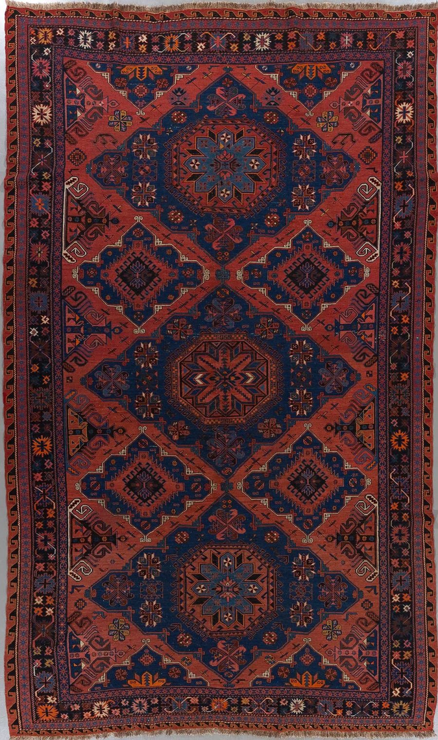 Traditioneller handgeknüpfter Teppich mit komplexem Muster in Rot-, Blau- und Beigetönen, Darstellung geometrischer Formen und floraler Motive, breiter Rand mit wiederholtem Muster und Fransen an den Enden.