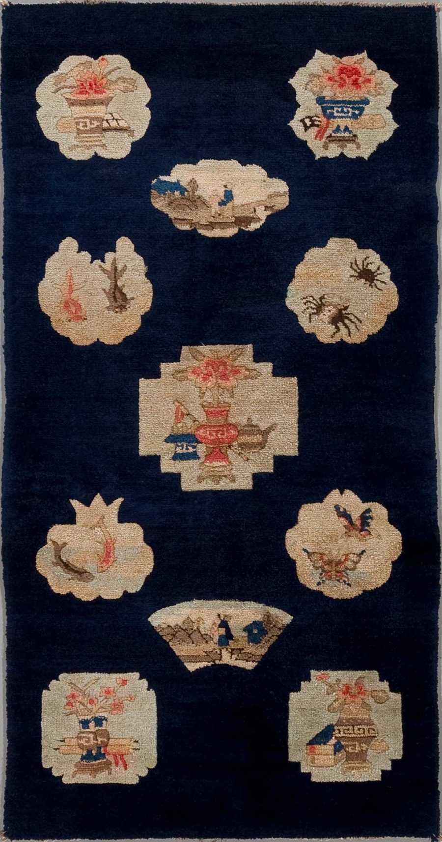 Antik wirkender Teppich mit dichtem Marineblaufeld und mehreren eingewebten traditionellen asiatischen Motiven in Beige- und Rottönen, darunter Vasen und Blumenarrangements, umgeben von einem schlichten, dunklen Rand.