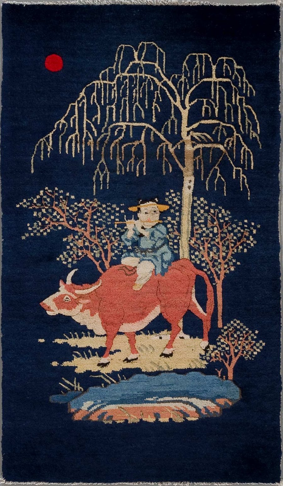 Alt Text: Textil-Kunstwerk mit stilisierter Darstellung eines Mädchens in blauer Kleidung, das auf einem roten Stier reitet, umgeben von einem großen weinenden Weidenbaum und weiteren Pflanzen, mit einem roten Sonnenpunkt im Hintergrund auf dunkelblauem Grund.