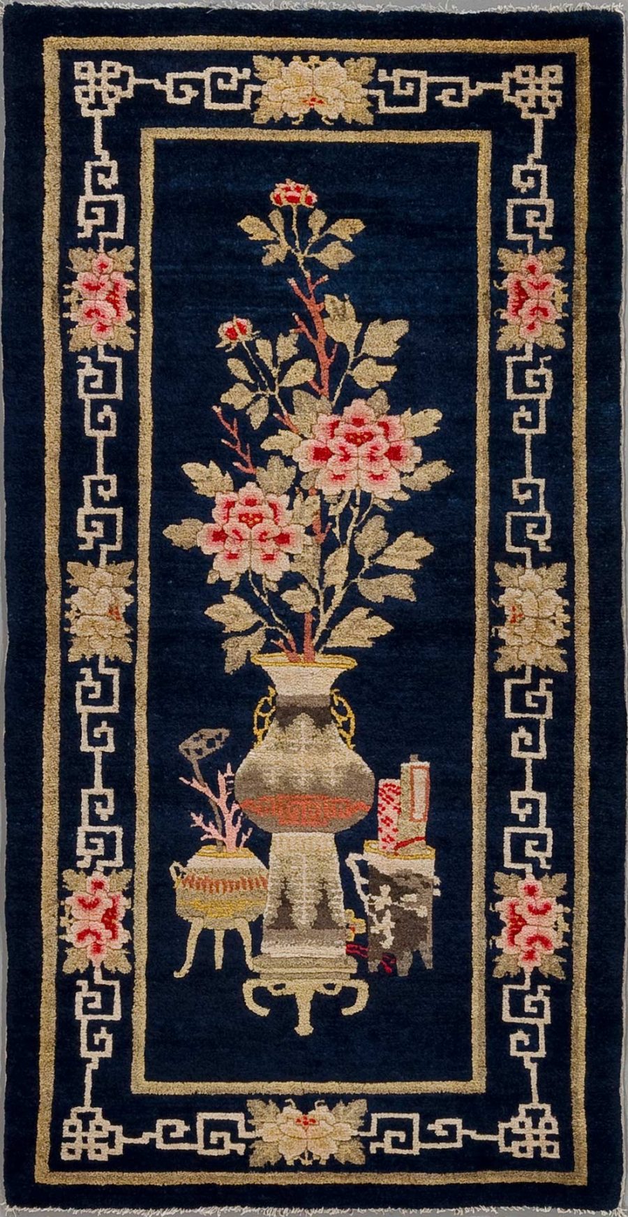 Orientteppich mit dunkelblauem Hintergrund und einem zentralen Motiv von Blumen in einer Vase, flankiert von traditionellen asiatischen Kunstgegenständen und umgeben von einer hellen Bordüre mit geometrisch-griechischem Muster.