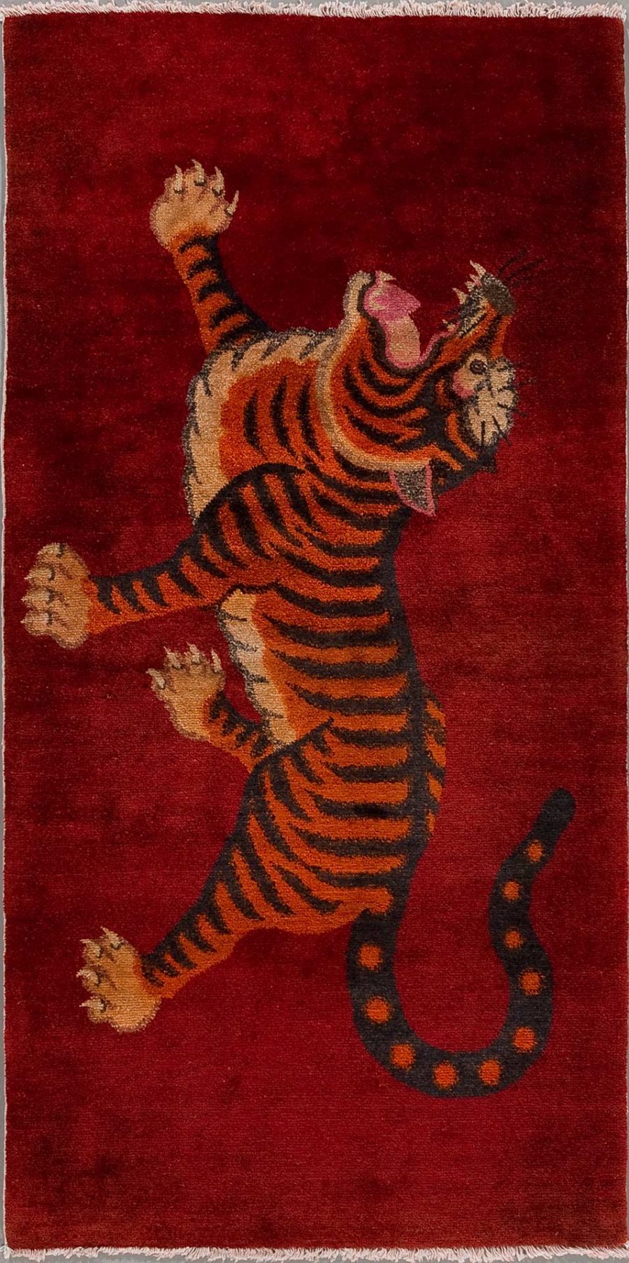 Handgeknüpfter Teppich mit einem aufrecht stehenden Tiger auf bordeauxrotem Hintergrund, der eine Pfote aggressiv erhebt und Mund und Augen in einem Ausdruck des Brüllens oder Fauchens öffnet.