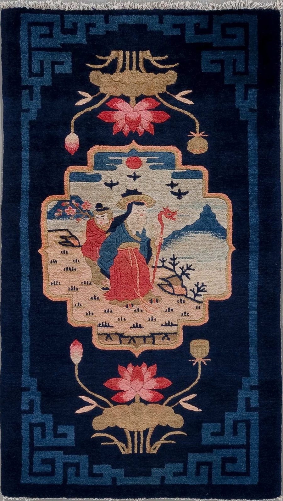 Traditioneller, vertikal orientierter, handgeknüpfter Teppich mit asiatisch anmutendem Design. Im Zentrum befindet sich eine Szene mit zwei stilisierten menschlichen Figuren, einer sitzend und einer stehend, umgeben von naturinspirierten Motiven wie Bäumen und Blumen. Oben und unten sind lotusähnliche Blumen abgebildet. Die Hauptfarben sind dunkelblau, beige, rot und Akzente in gedeckten Farbtönen, umrandet von einer Bordüre mit geometrischem Muster.