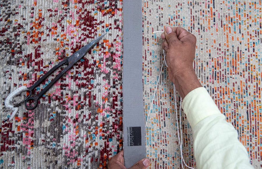 Handwerker bei der Arbeit an einem Teppich mit buntem Muster, daneben eine geöffnete Schere und Kantenband mit einem Logo.