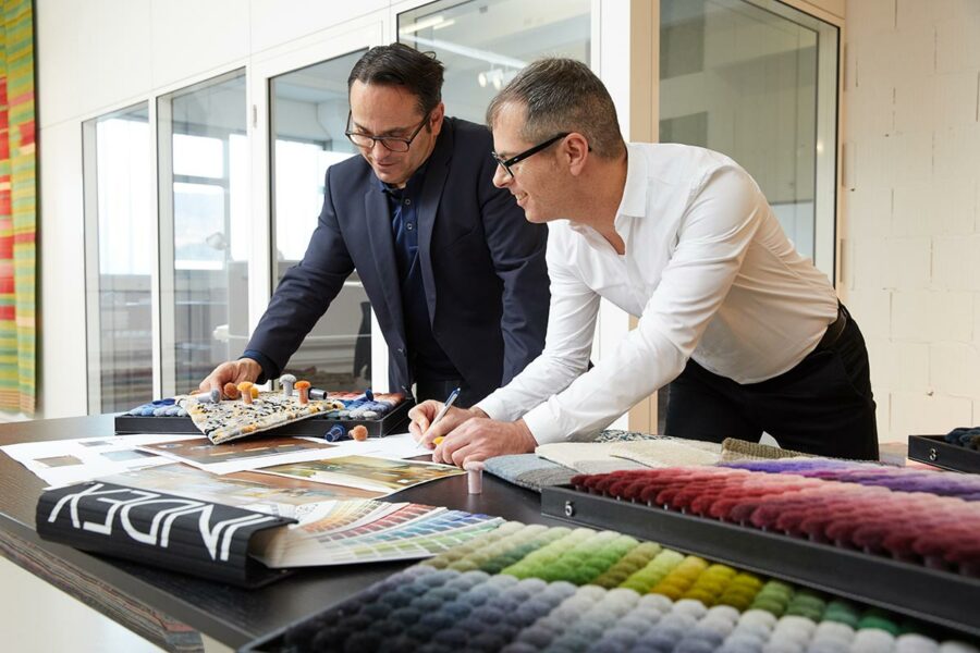Zwei Männer in Business-Kleidung betrachten und diskutieren Materialproben und Farbpaletten auf einem Tisch in einem hellen Büro.