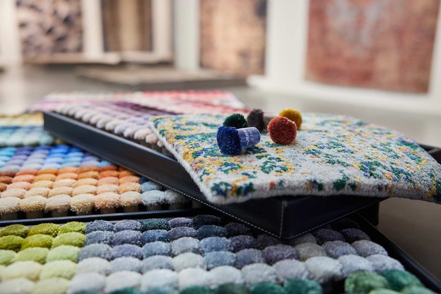 Ein Musterbuch für Teppiche mit vielen verschiedenen Farb- und Texturoptionen, die zur Auswahl auf mehreren offenen Seiten präsentiert werden, mit einem Fokus auf einem Muster mit bunten Blumen auf der oberen Seite. Im Hintergrund unscharfe Kunstwerke an einer Wand.