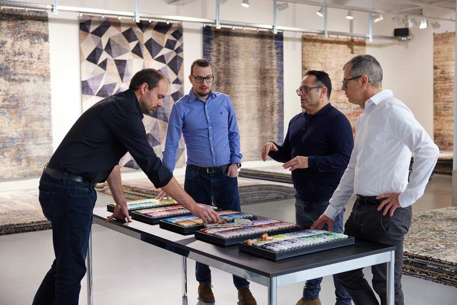Vier Männer betrachten und diskutieren über Farbmuster auf einer Tischplatte in einem hell beleuchteten Ausstellungsraum mit verschiedenen Teppichen im Hintergrund.
