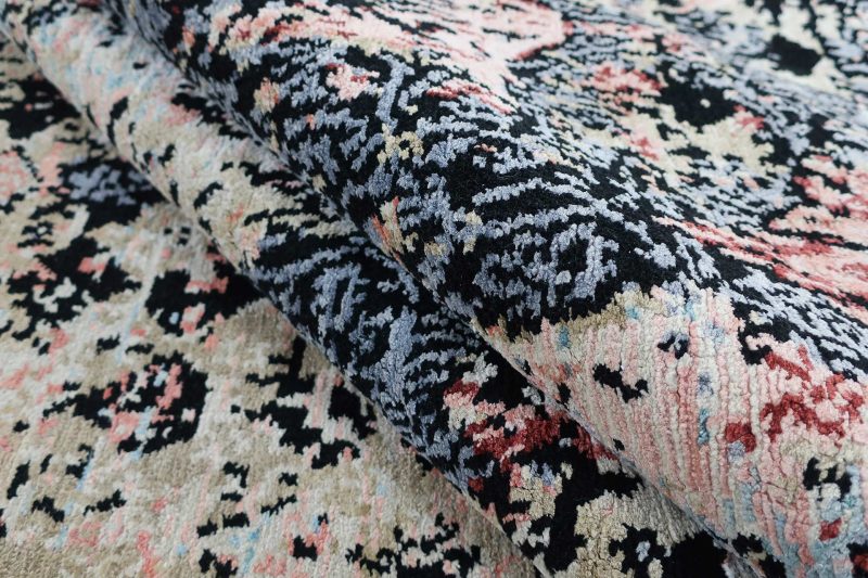 Nahaufnahme eines gefalteten, gemusterten Teppichs mit komplexen, traditionellen Designs in Schwarz, Blau, Rosa und Beige.
