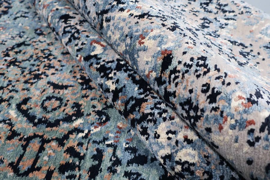Detailansicht eines gemusterten Teppichs mit gefalteter Ecke, das Muster zeigt abstrakte Formen in Blau, Schwarz, Orange und Beige auf hellem Grund.