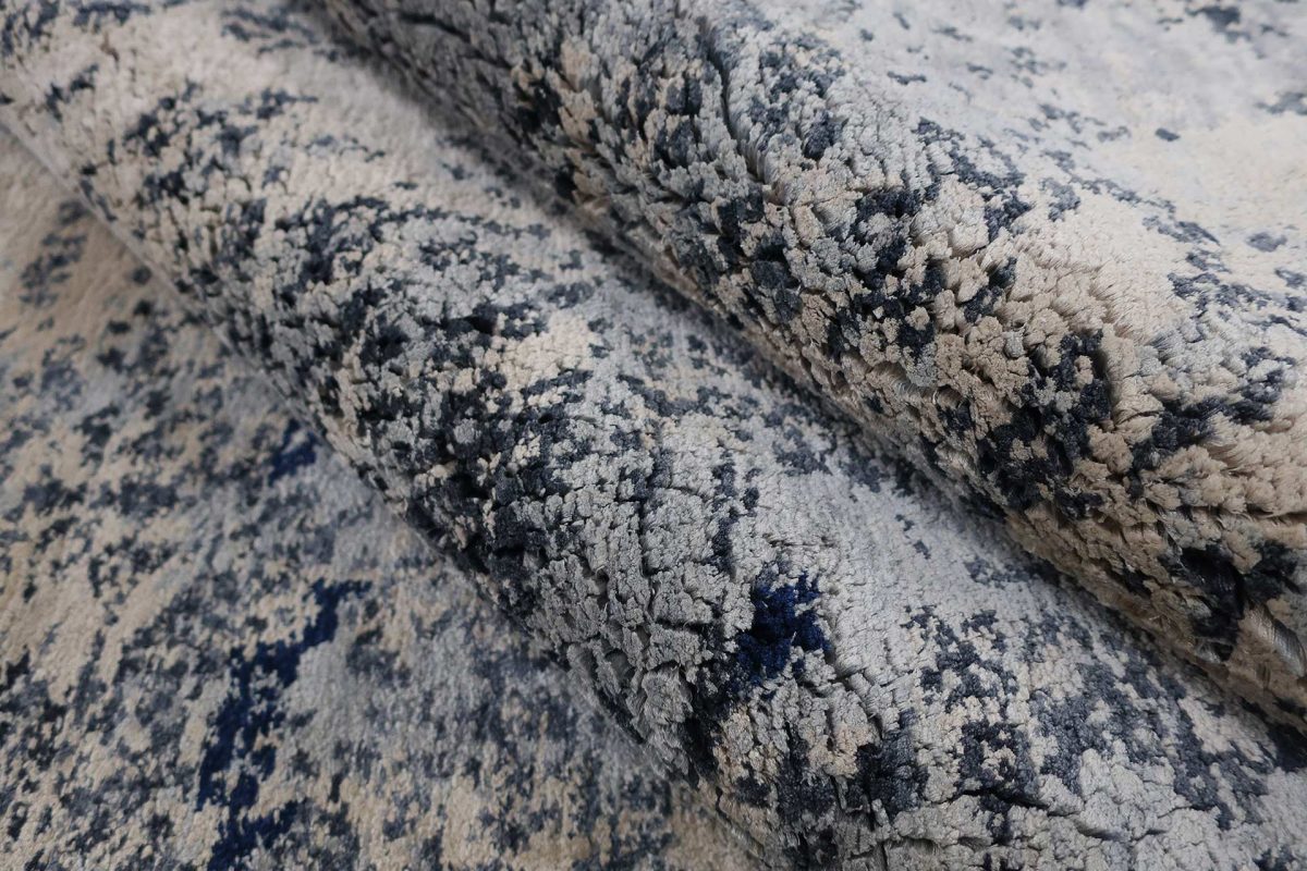 Nahaufnahme eines gemusterten Teppichs in Grau- und Beigetönen mit texturierter Oberfläche, zusammengerollt im Vordergrund.