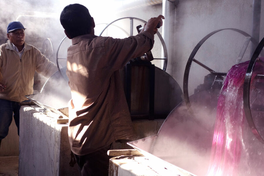 Zwei Männer arbeiten in einer Weinfabrik mit großen Metallbehältern, einer bedient einen Hahn, aus dem ein purpurroter Flüssigkeitsstrahl fließt, während Dampf in die Luft steigt.