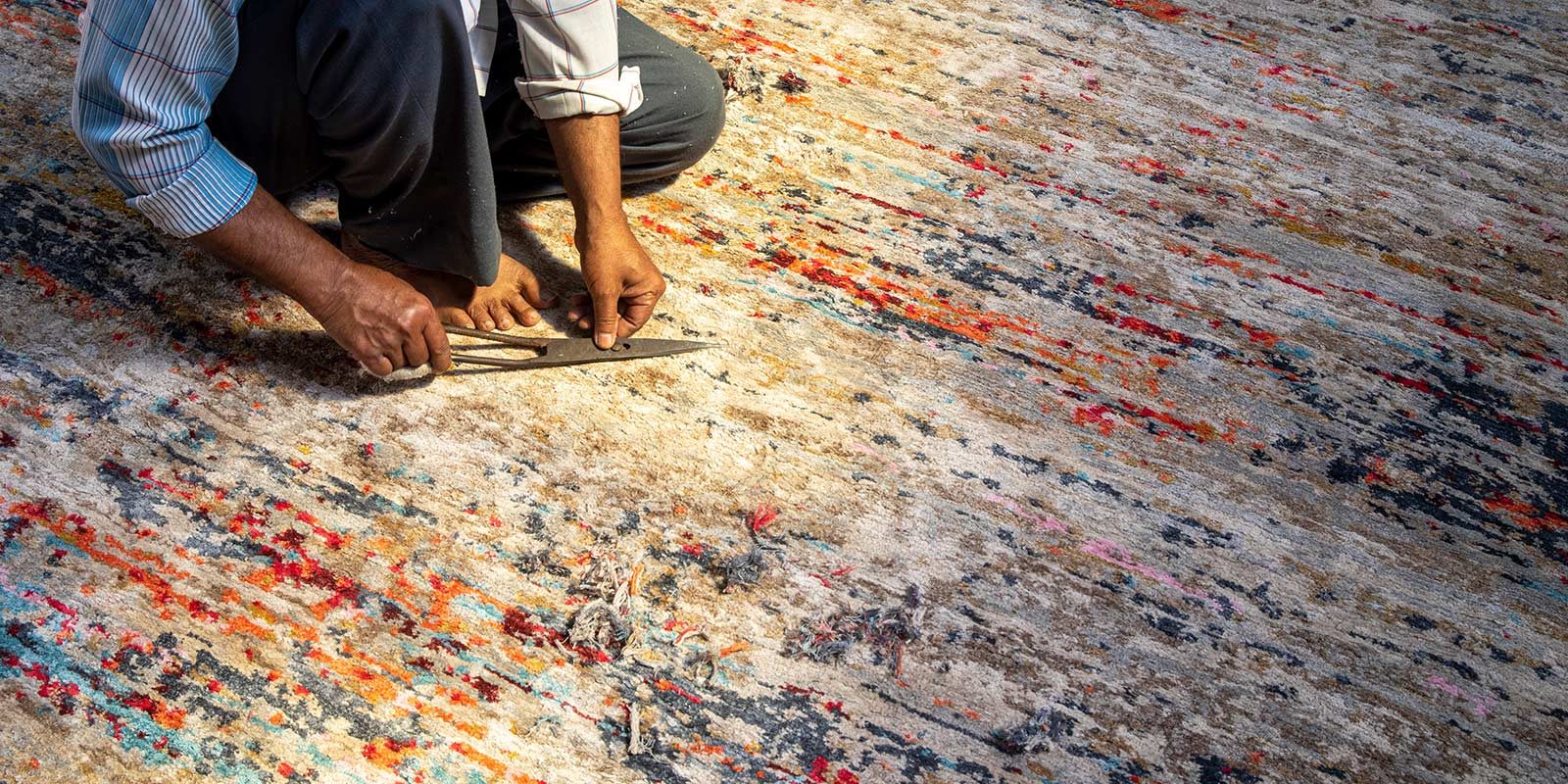 Person in halber Draufsicht, die auf dem Boden kniet und mit einer Schere Stoff schneidet; bunter Teppich mit abstraktem Muster im Hintergrund.