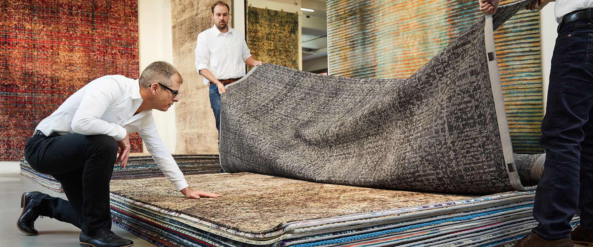 Zwei Männer in einem Teppichgeschäft beim Umschichten von Teppichen auf einem großen Stapel, mit verschiedenen Teppichdesigns im Hintergrund sichtbar.
