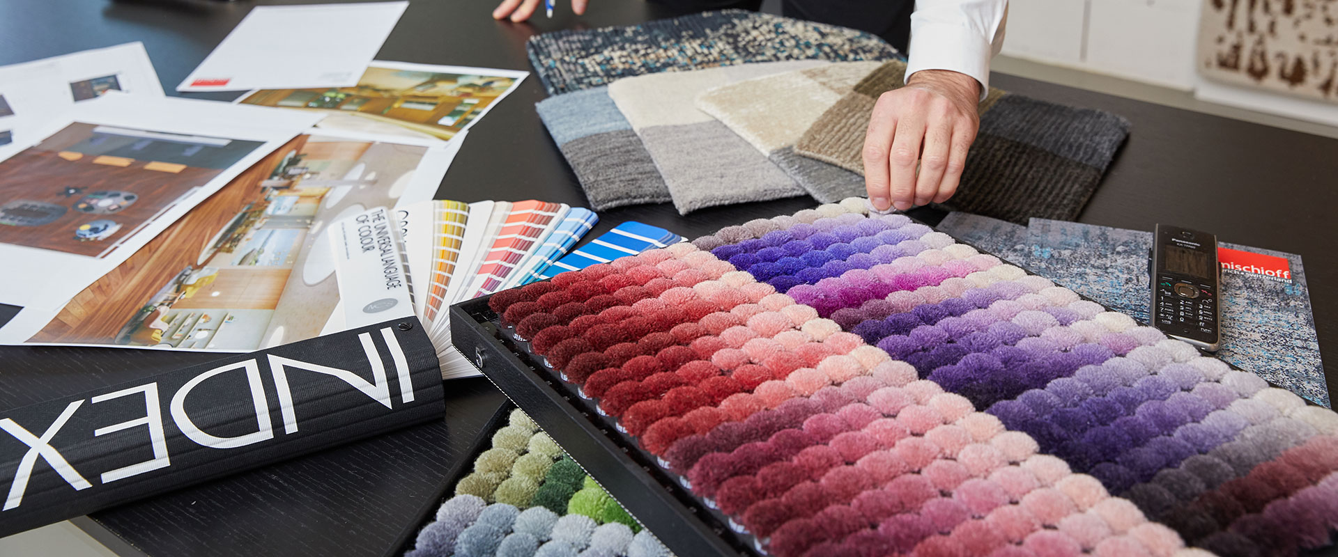 Innenarchitektur-Arbeitsplatz mit Farbfächern, Textilmustern, Bodenbelag-Proben und inspirierenden Interieurfotos auf einem Tisch, Hand einer Person berührt Teppichmuster.