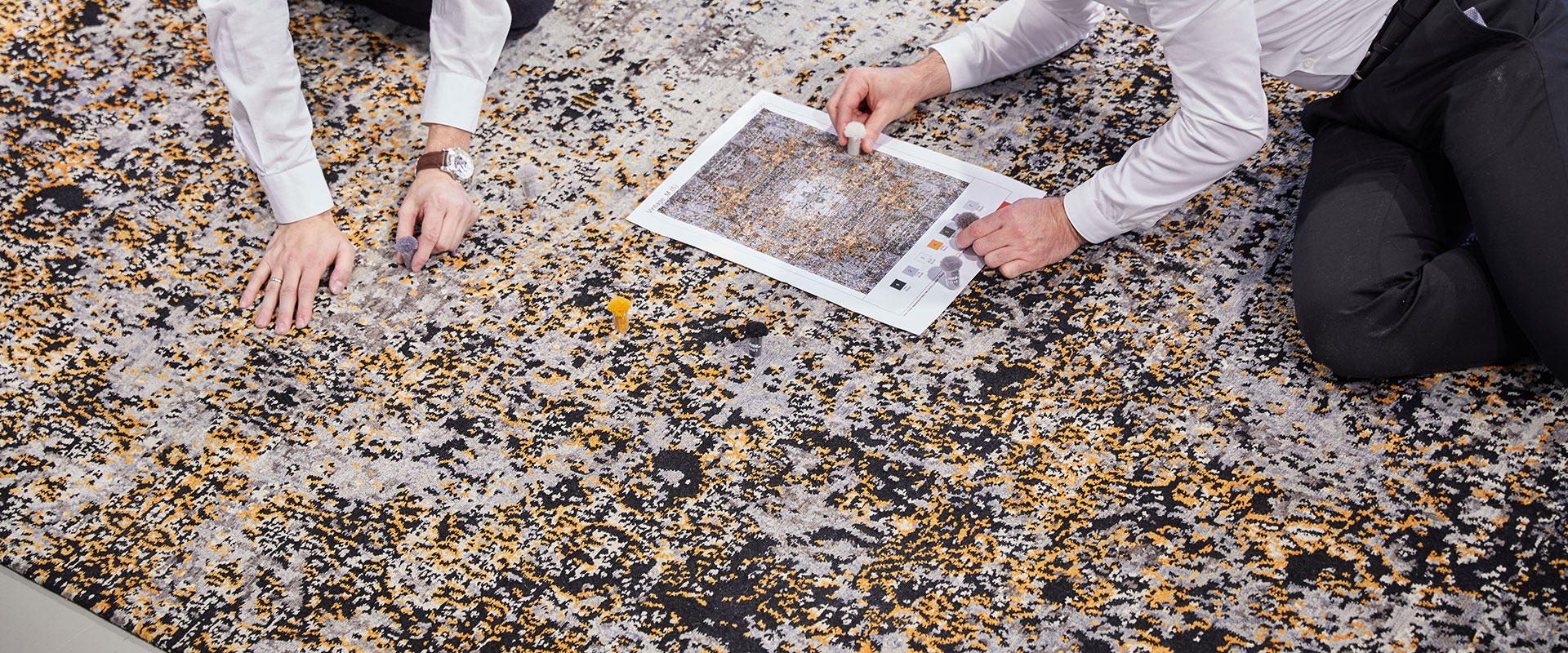 Zwei Personen knien auf einem gemusterten Teppich und untersuchen ein Puzzle; eines der Puzzleteile liegt neben ihnen. Sie halten ein Referenzbild des Puzzle-Designs. Der Fokus liegt auf den Händen und Puzzleteilen, Gesichter sind nicht sichtbar.