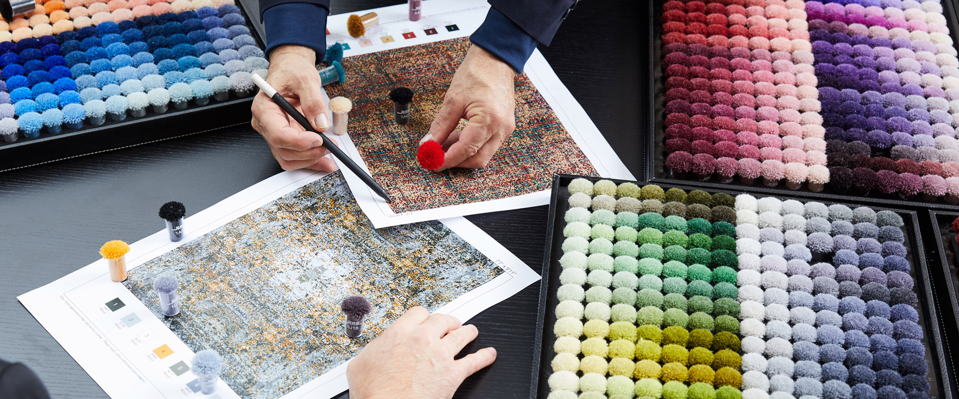 Designprofis betrachten und wählen Teppichmuster und -farben aus, dargestellt durch farblich markierte Pompoms auf Farbkarten und Musterbüchern, auf einem dunklen Tisch mit mehreren Farbplatten und Textilproben im Fokus.